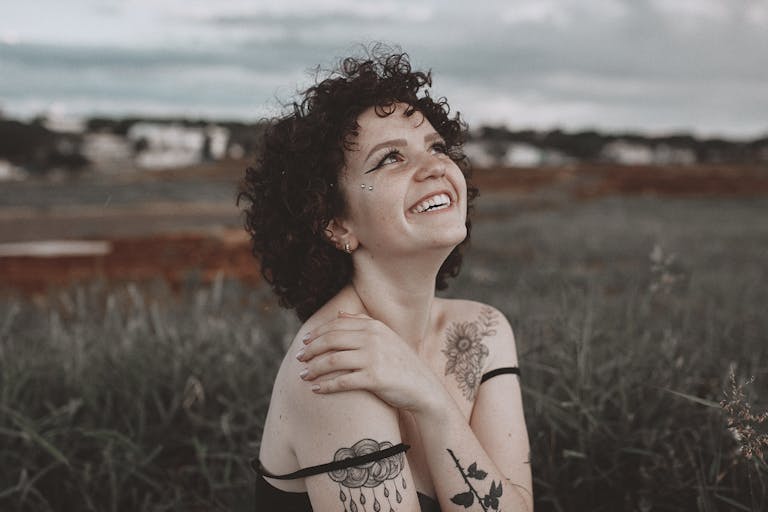 Glimmers in Spirituality: Tiny Moments That Raise Your Energy and Vibration - A smiling woman with tattoos enjoys the outdoors