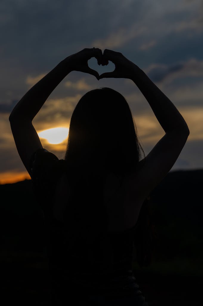 Silhouette of a womans shadow forming a heart shape with hands at sunset, symbolizing love and peace.
