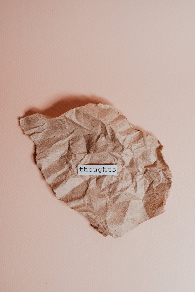 A crumpled brown paper on a pink background with the word 'thoughts' in focus to represent negative thoughts and inner critic.