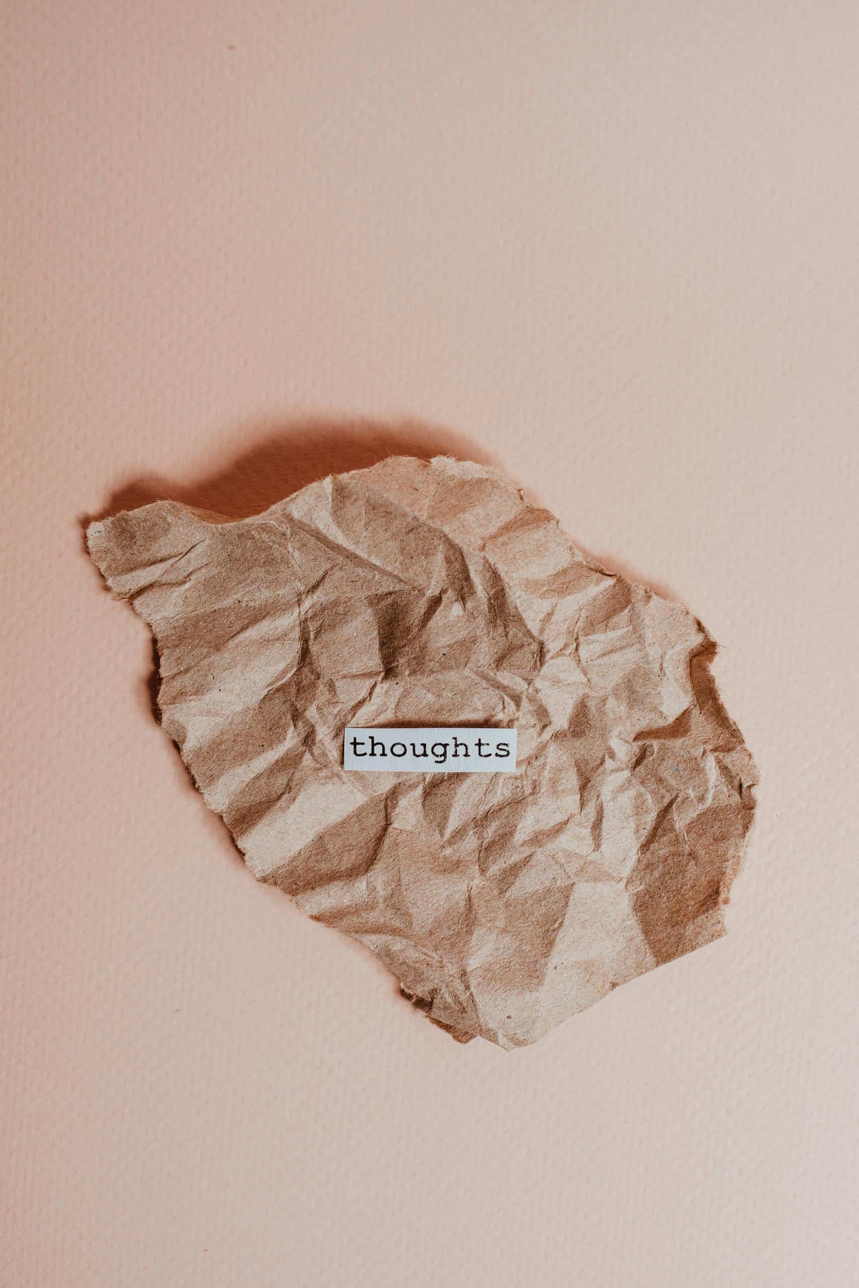 A crumpled brown paper on a pink background with the word 'thoughts' in focus to represent negative thoughts and inner critic.